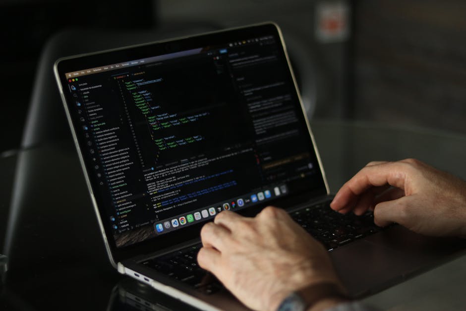 A close-up of a person's hand typing on a laptop keyboard placed on a wooden desk, with visible code on the screen. The person wears a wristwatch, and the background is plain and out of focus. The set